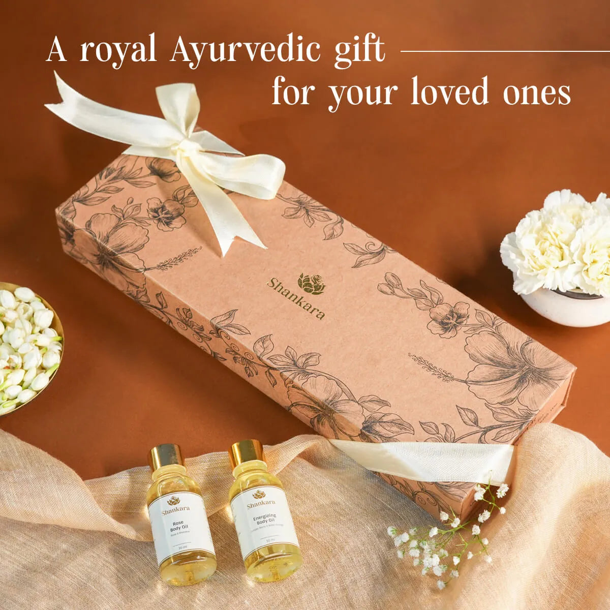 A Shankara Ayurvedic gift set featuring a brown patterned box tied with a cream ribbon, alongside two small golden Ayurvedic oil bottles on a beige fabric surface, surrounded by white flowers on a warm brown background.