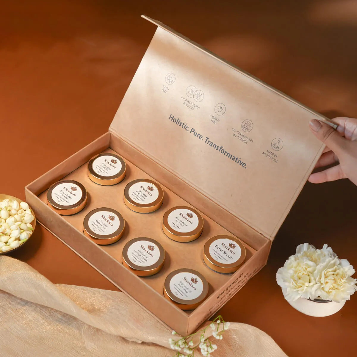 shankara forest bloom renew glow gift box containing eight small round jars with gold lids and white labels, arranged on a warm brown surface. A hand is holding the open lid, with decorative flowers and draped fabric around the box.
